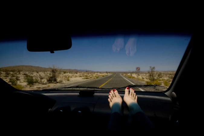 Feet with red nail polish resting on a car dashboard facing an empty desert highway