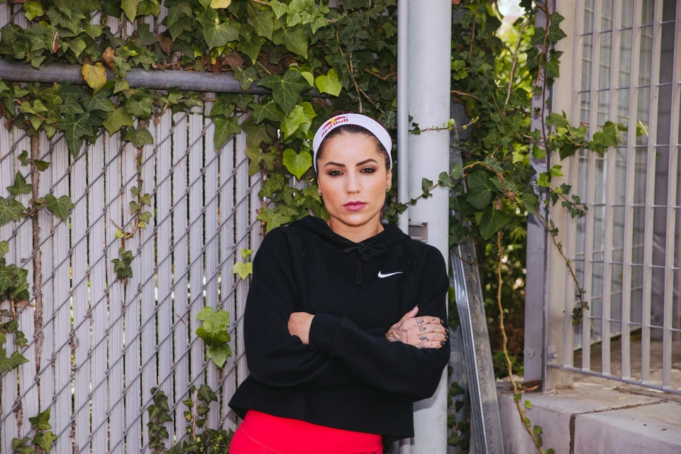 Adult woman in athletic attire poses outdoors against a fence covered in vines. She wears a Red Bull headband and a black Nike hoodie.