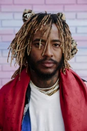 Portrait of a man with dreadlocks, wearing a red scarf, white t-shirt, and pearl necklaces against a pink brick wall.