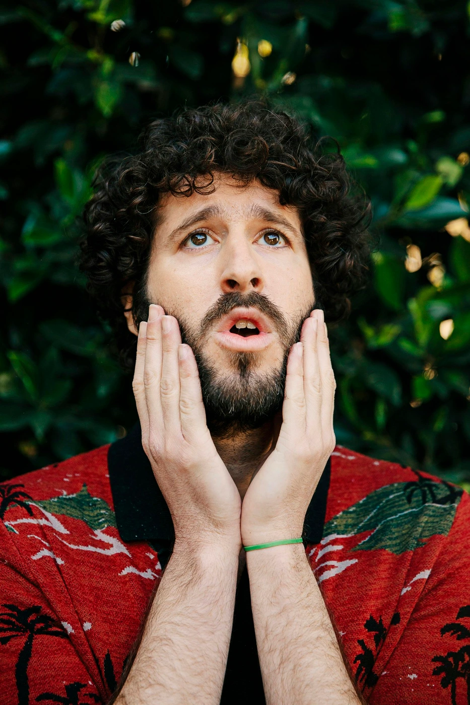 Man with curly brown hair and beard holding his hands to his face in surprise against a green foliage background