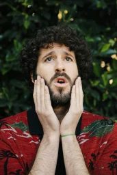 Man with curly brown hair and beard holding his hands to his face in surprise against a green foliage background