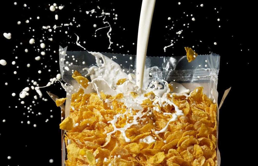 Golden corn flakes cereal in a clear plastic bag with fresh milk pouring, creating splashes against a black background.