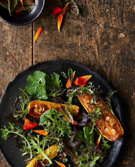 Overhead shot of a black plate with a butternut squash salad with greens and petals on a rustic wooden table; culinary photography.