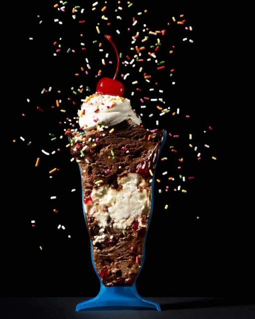 An ice cream sundae in a blue glass is decorated with a cherry on top and colorful sprinkles against a dark background.