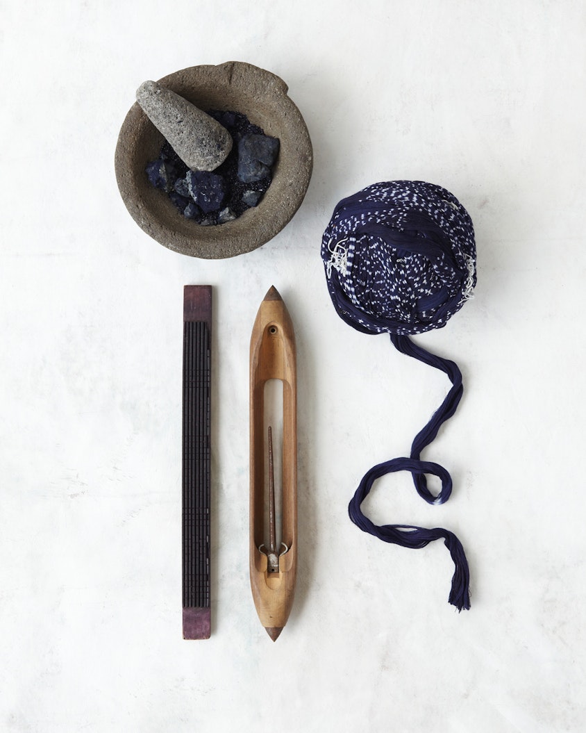 Flat lay of a stone mortar and pestle with blue dye chunks, weaving tools, and tied blue fabric on a white marble surface.