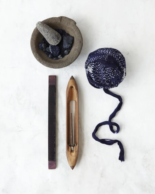 Flat lay of a stone mortar and pestle with blue dye chunks, weaving tools, and tied blue fabric on a white marble surface.