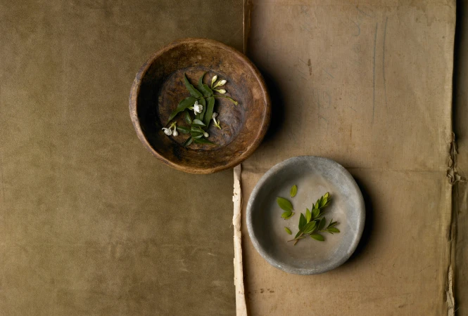 Top-down view of two rustic bowls containing green leaves and white flowers resting on textured parchment and olive fabric.