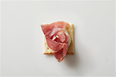 Overhead shot of cracker topped with a small scoop of mozzarella cheese and prosciutto, food photography, white background.