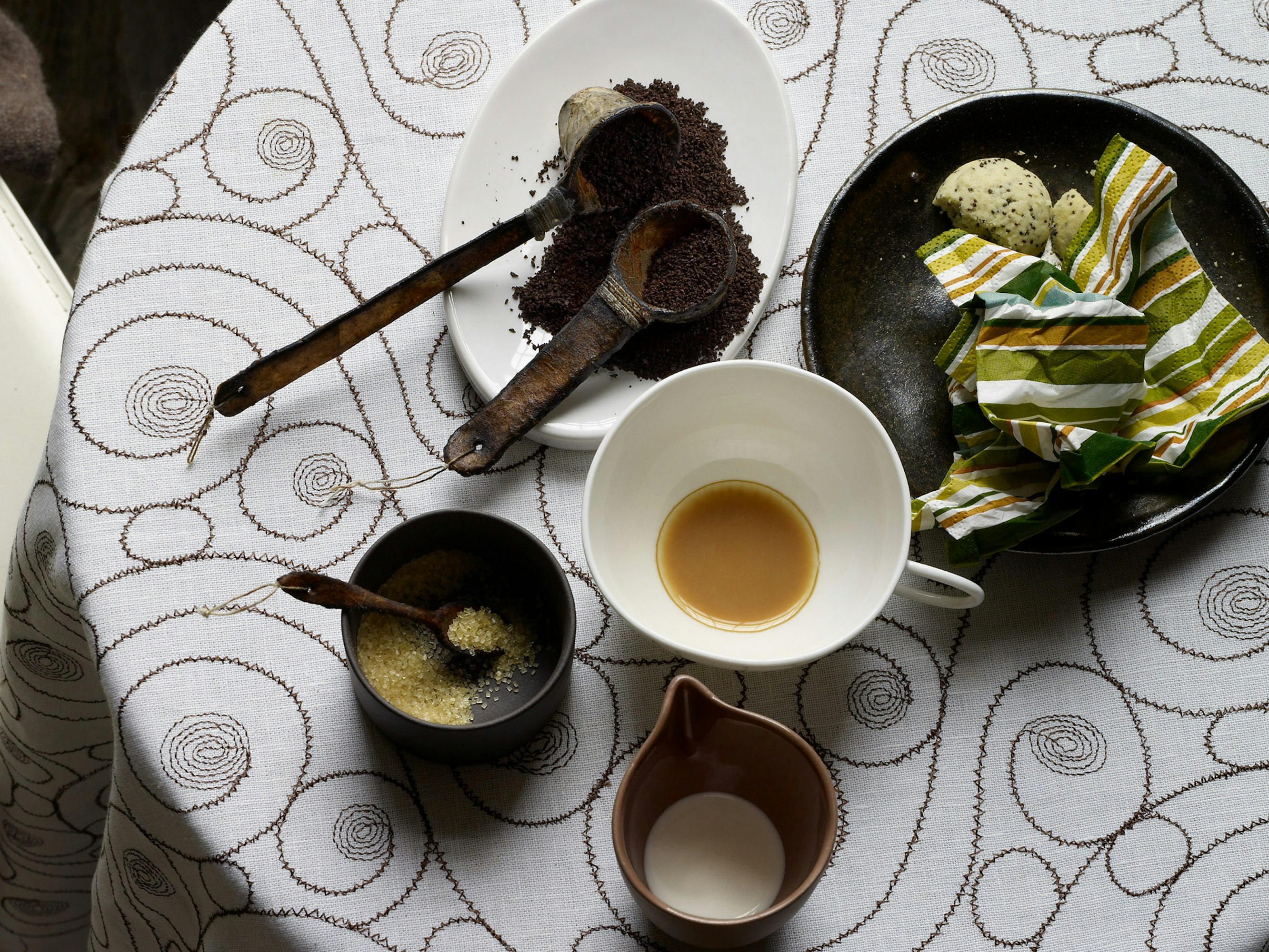 Overhead shot of coffee and tea service with wooden spoons, cookies, and a striped napkin on a decorative white tablecloth.
