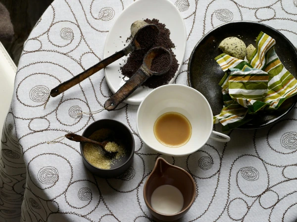 Overhead shot of coffee and tea service with wooden spoons, cookies, and a striped napkin on a decorative white tablecloth.