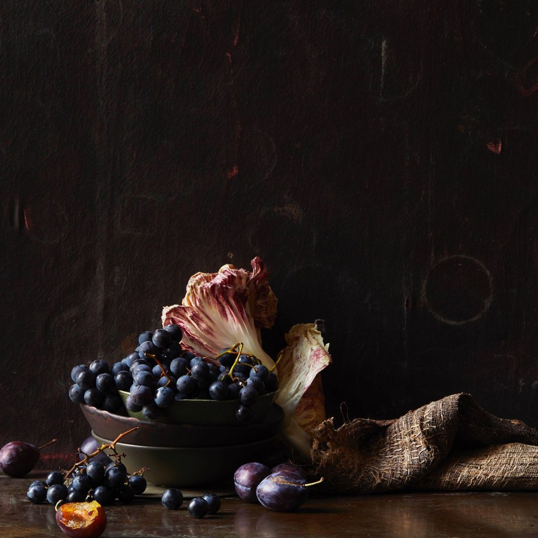 Dark still life: black grapes cascading from bowls with plum fruits, radicchio leaf, and burlap sack on a wood table against a dark backdrop.