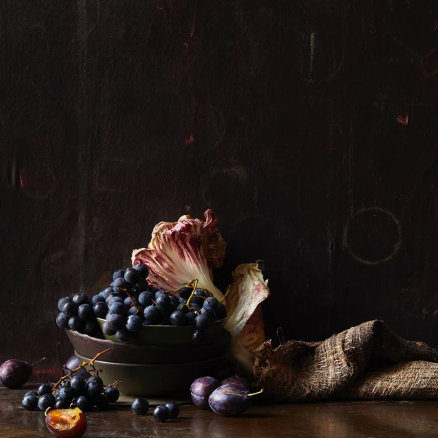 Dark still life: black grapes cascading from bowls with plum fruits, radicchio leaf, and burlap sack on a wood table against a dark backdrop.
