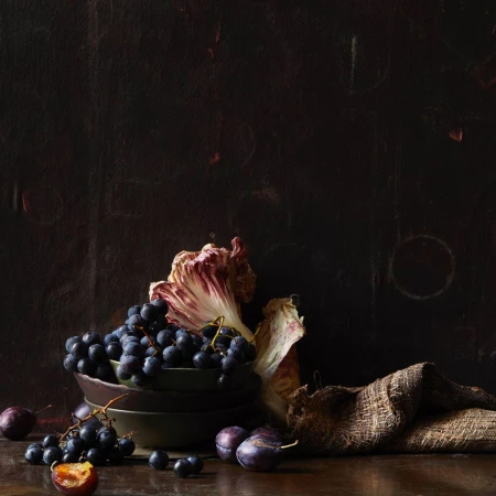 Dark still life: black grapes cascading from bowls with plum fruits, radicchio leaf, and burlap sack on a wood table against a dark backdrop.