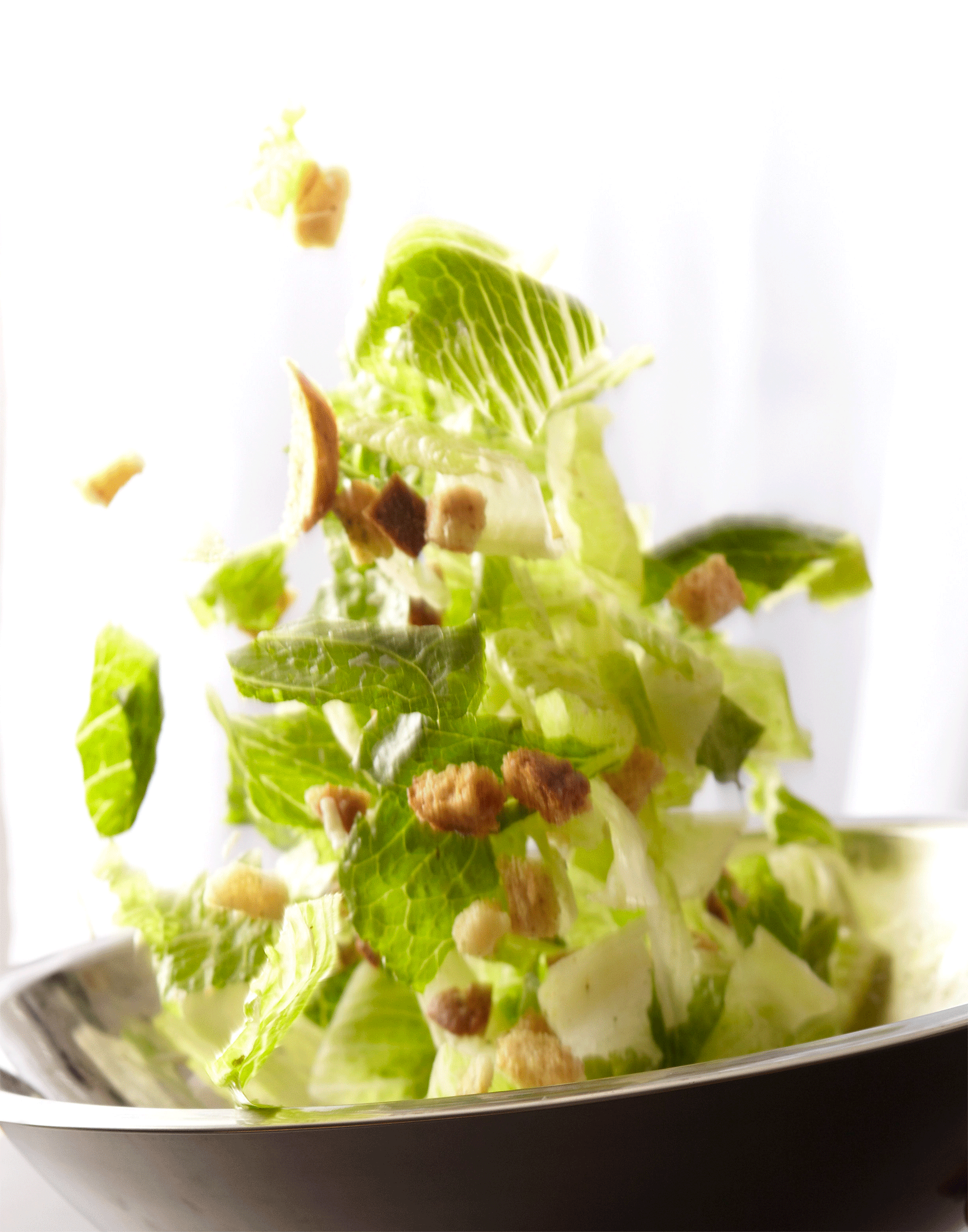 Close-up of caesar salad with romaine lettuce and croutons falling into a metal bowl against a bright white background in a studio.