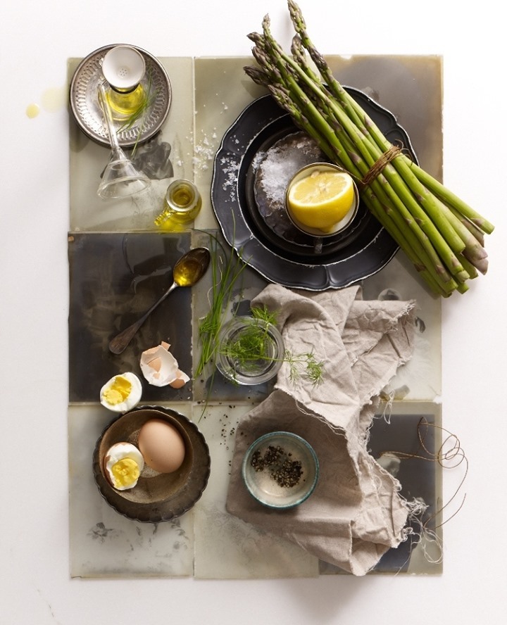 Overhead shot of asparagus tied with twine, halved lemon, oil, herbs, and halved boiled eggs arranged on tiled surface.