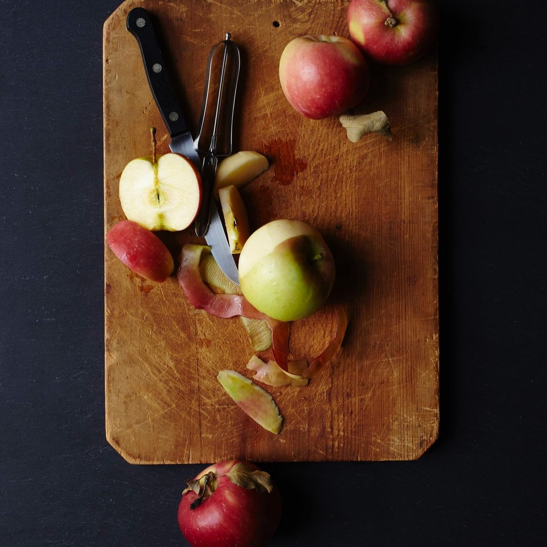 Overhead shot of a wooden cutting board with whole and sliced apples, a peeler, and a knife against a dark background. Food photography.