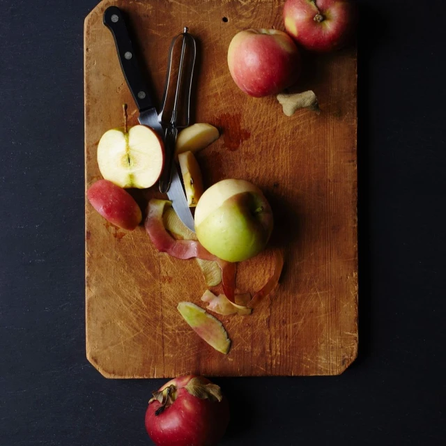 Overhead shot of a wooden cutting board with whole and sliced apples, a peeler, and a knife against a dark background. Food photography.