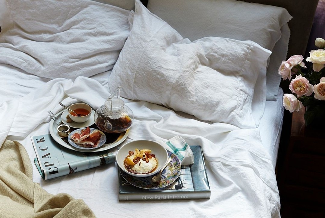 Breakfast in bed features a tray with tea, fruit, and pastries on white linen bedding with roses in a bedroom setting.