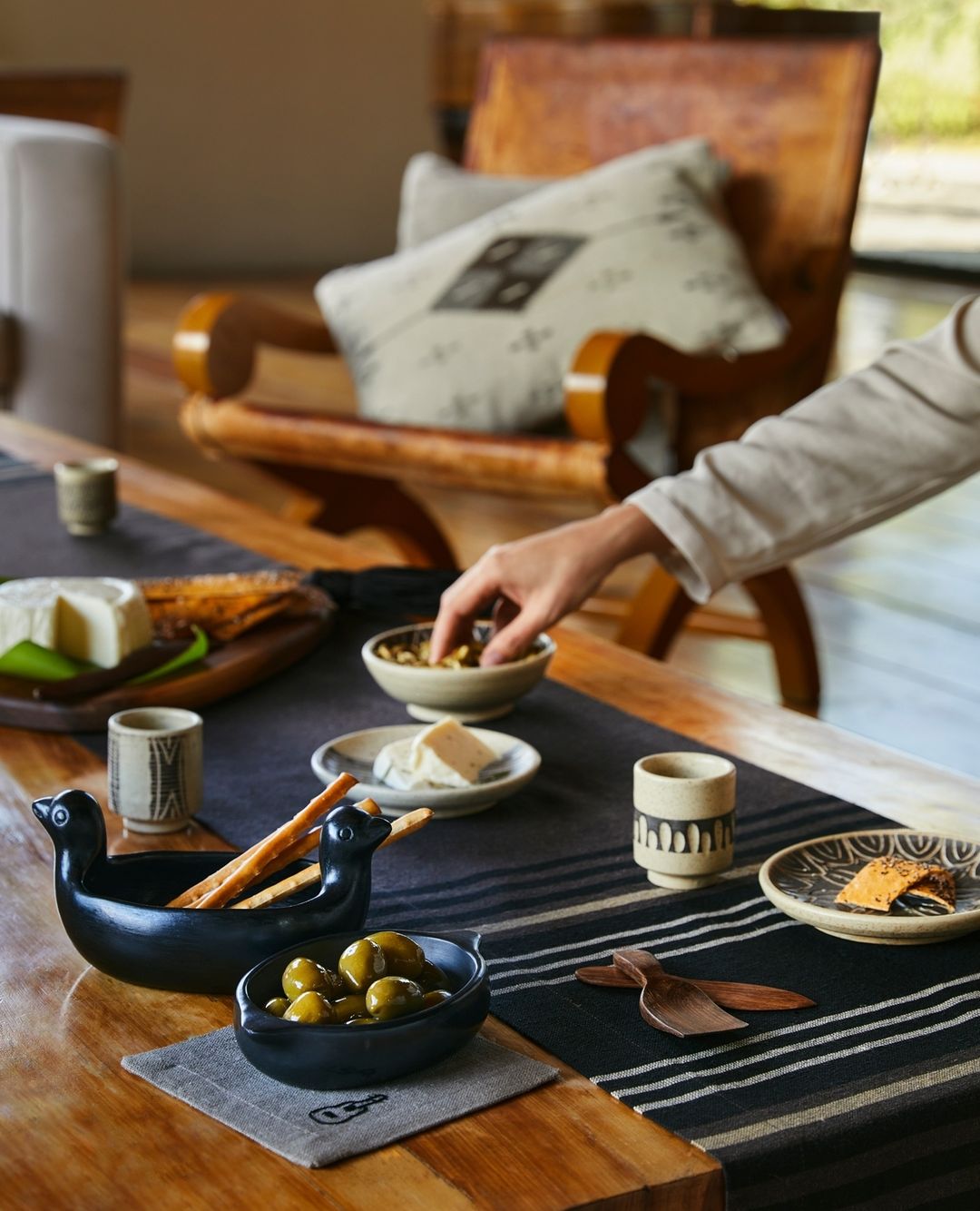 Overhead shot of a wooden table set with artisan cheese, olives, ceramic dishes, wooden utensils, and a neutral-toned table runner.