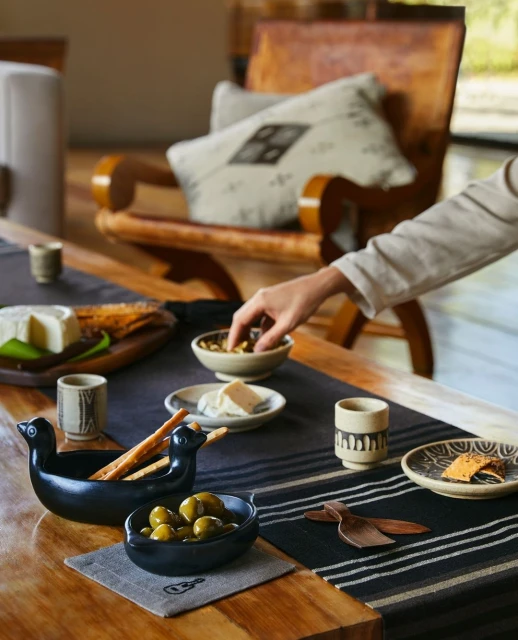 Overhead shot of a wooden table set with artisan cheese, olives, ceramic dishes, wooden utensils, and a neutral-toned table runner.