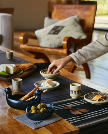 Overhead shot of a wooden table set with artisan cheese, olives, ceramic dishes, wooden utensils, and a neutral-toned table runner.
