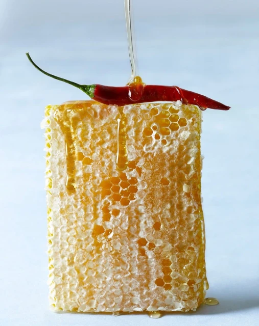 Honeycomb block with a red chili pepper on top, honey being poured over, against a light blue background. Food and drink photography.
