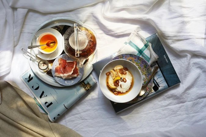 Overhead shot of tea service, jamón, and rice bowl on bed with white sheets and books suggesting a cozy, relaxed morning.