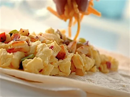 Close-up of a person sprinkling shredded cheddar cheese over a burrito filling of scrambled eggs, potatoes, peppers, and onions.