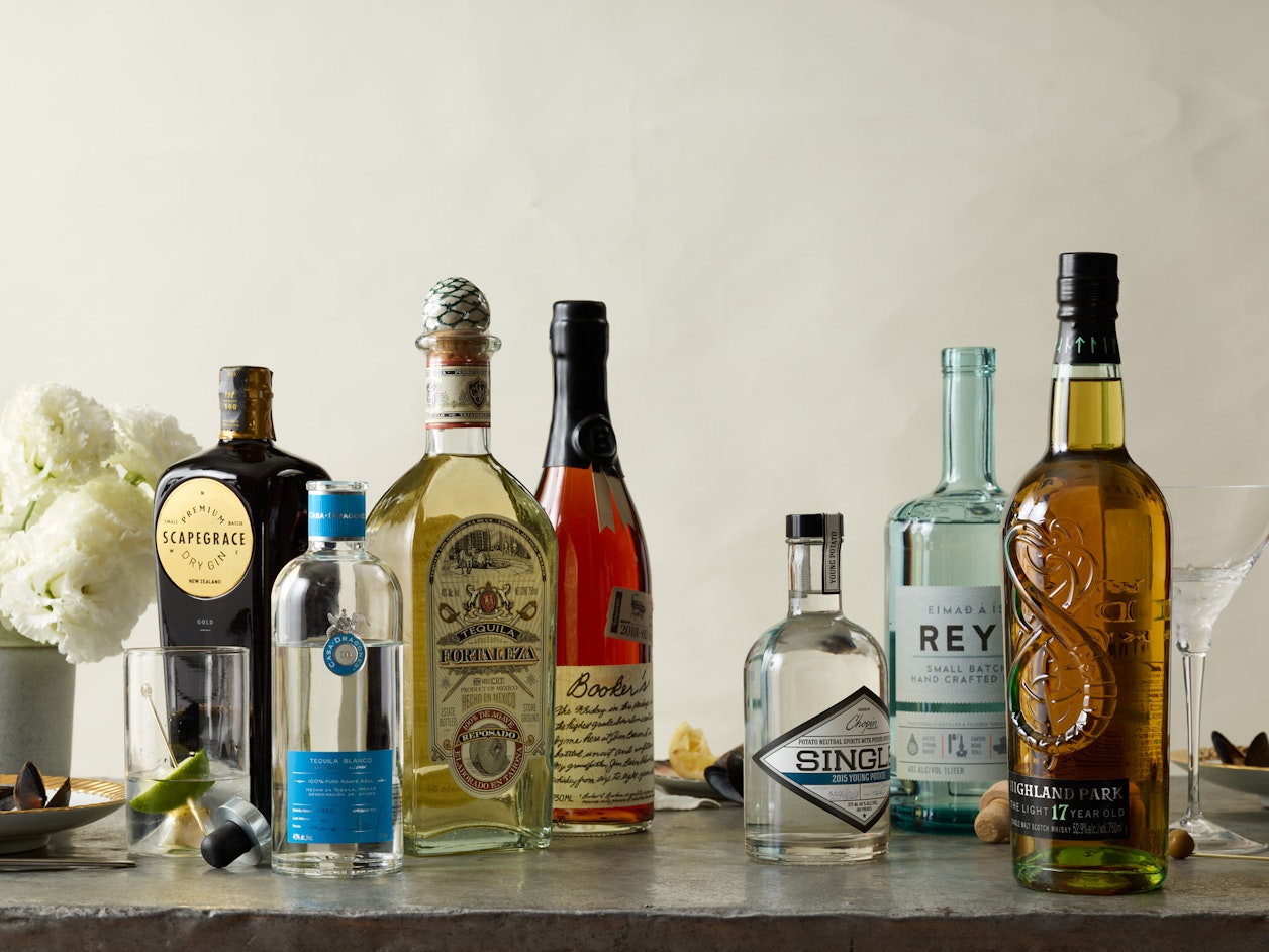 Arrangement of various spirit bottles including gin, tequila, whiskey and cocktails on a gray countertop with white flowers in background.
