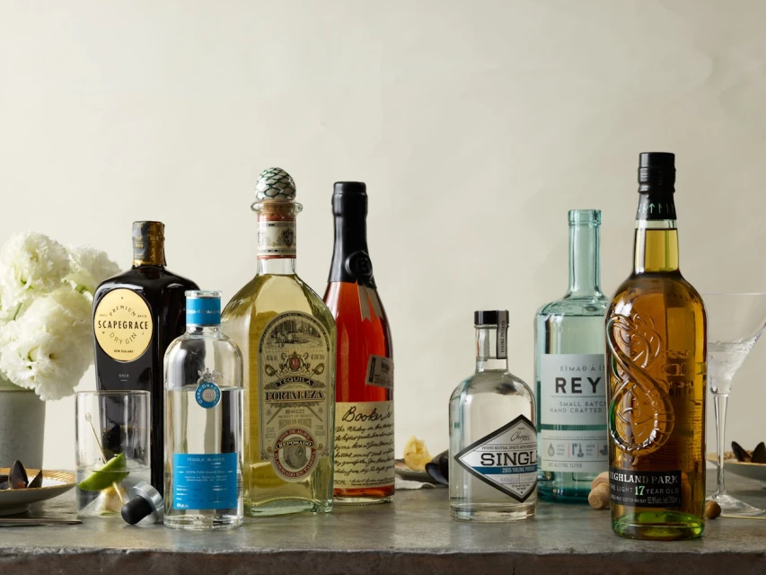 Arrangement of various spirit bottles including gin, tequila, whiskey and cocktails on a gray countertop with white flowers in background.