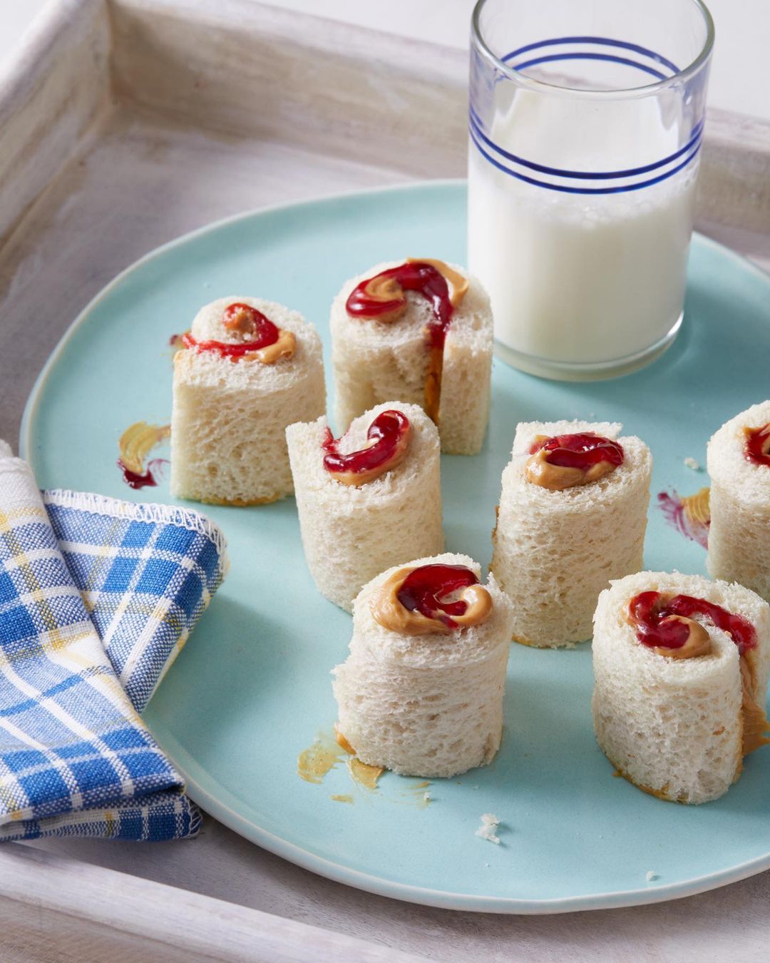 Several peanut butter and jelly roll sandwiches on a light-blue plate with a glass of milk and blue-checkered napkin on a white tray.