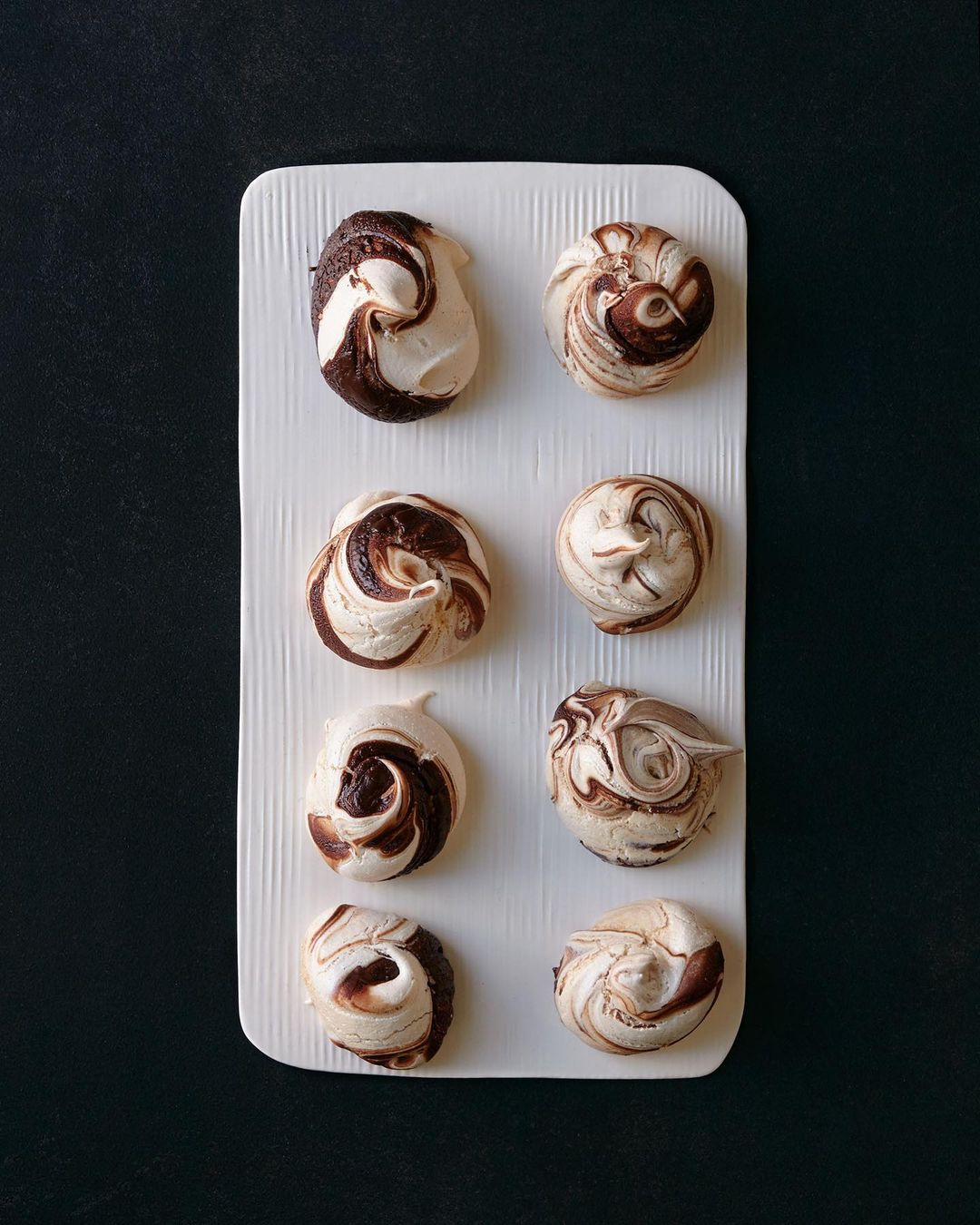 Overhead shot of eight swirled chocolate and vanilla meringues arranged on a white rectangular plate against a dark background.