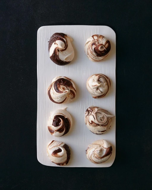 Overhead shot of eight swirled chocolate and vanilla meringues arranged on a white rectangular plate against a dark background.
