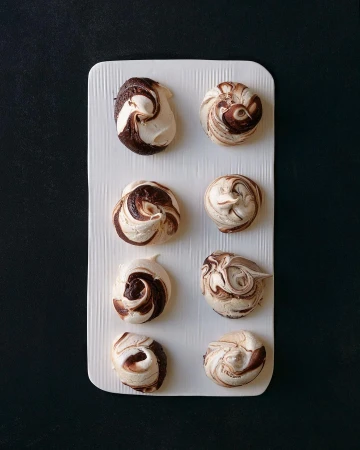 Overhead shot of eight swirled chocolate and vanilla meringues arranged on a white rectangular plate against a dark background.