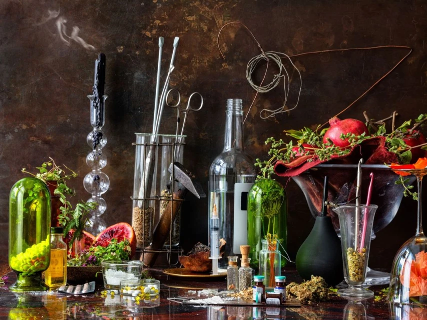 Dramatic still life features assorted glass bottles and vessels filled with liquids, dried herbs, and seeds arranged on a dark surface.