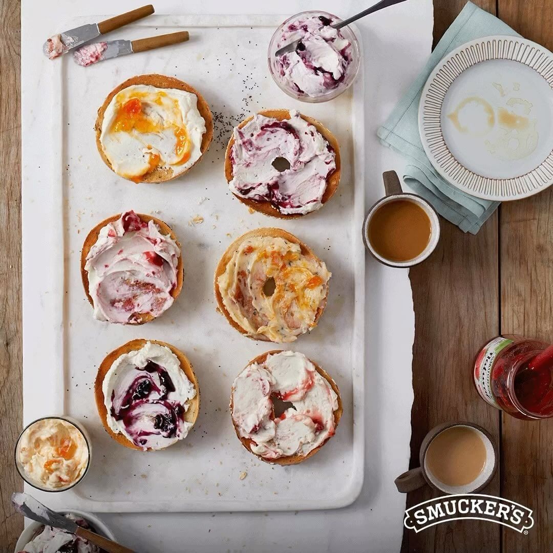 Overhead shot of bagels topped with various cream cheese and fruit spread combinations, served on a white tray with coffee mugs.