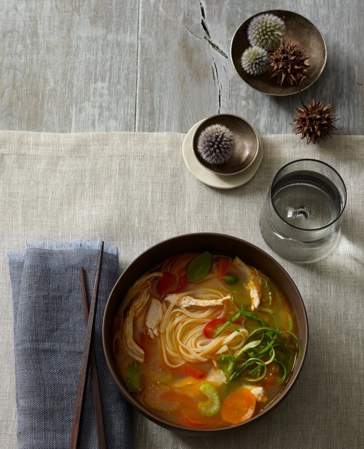 Overhead shot of chicken noodle soup in a dark bowl next to a linen napkin and a glass of water on a gray surface.