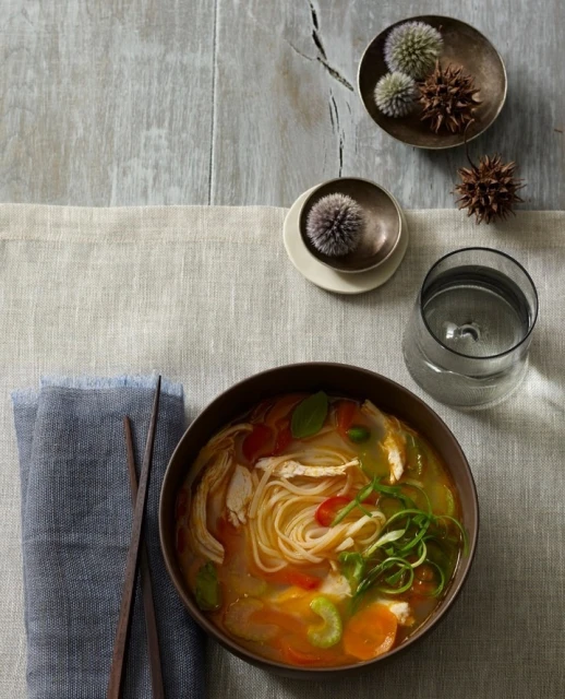 Overhead shot of chicken noodle soup in a dark bowl next to a linen napkin and a glass of water on a gray surface.