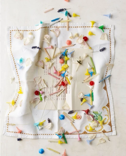 Overhead shot of a decorated white linen with candles, beads, flower petals, and sticks scattered on top against a white backdrop.