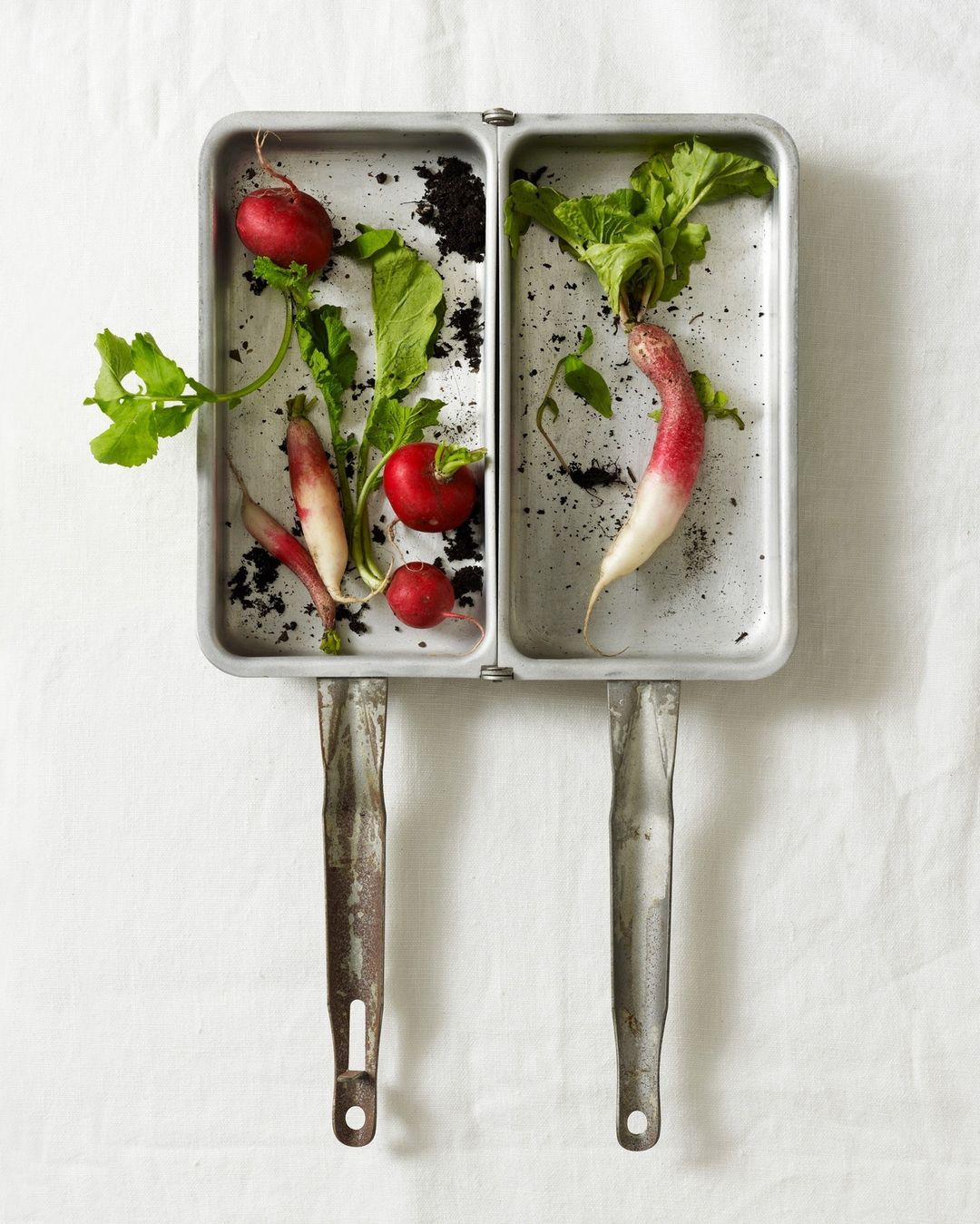 Overhead shot of fresh radishes with green leaves inside a metal tray with rustic handles, set against a textured white cloth.