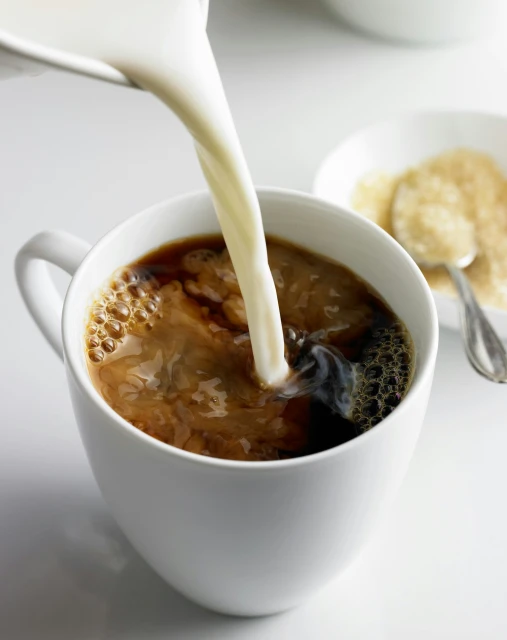 Creamy milk is poured into a white mug filled with rich black coffee, alongside a bowl of sugar, creating a warm, inviting beverage.