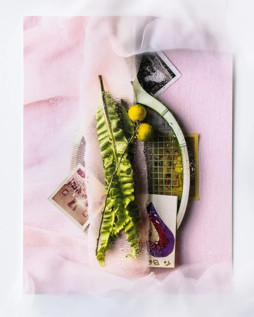 Still life of tennis racket, leaves, and photos on pink cloth backdrop creating a visually interesting and artistic composition.