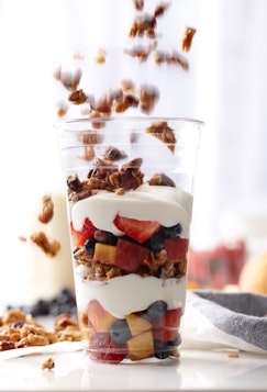 A clear plastic cup filled with layers of yogurt, granola, and fresh fruit like strawberries and blueberries, with granola falling from above.