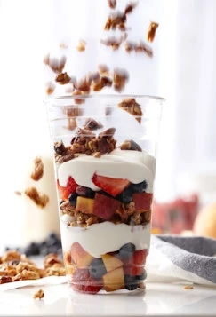 A clear plastic cup filled with layers of yogurt, granola, and fresh fruit like strawberries and blueberries, with granola falling from above.