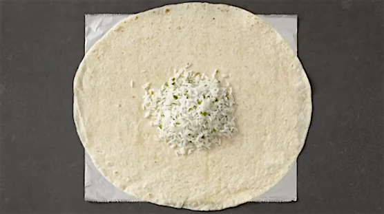 Top-down view of a large flour tortilla filled with a mound of white rice and herbs on a white parchment paper and dark gray backdrop.