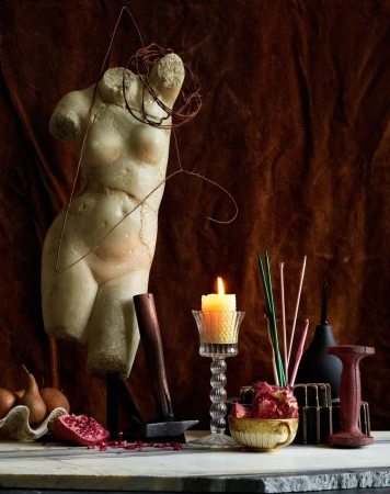 Still life of a female torso statue, candle, fruit, and other objects arranged on a marble surface against a brown fabric backdrop.