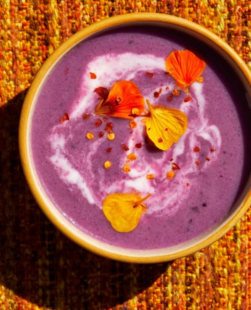 Overhead view of a bowl filled with purple cream soup garnished with swirls of white cream, edible orange and yellow flower petals, and chili flakes.