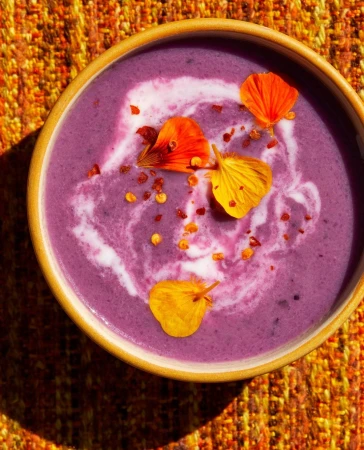 Overhead view of a bowl filled with purple cream soup garnished with swirls of white cream, edible orange and yellow flower petals, and chili flakes.
