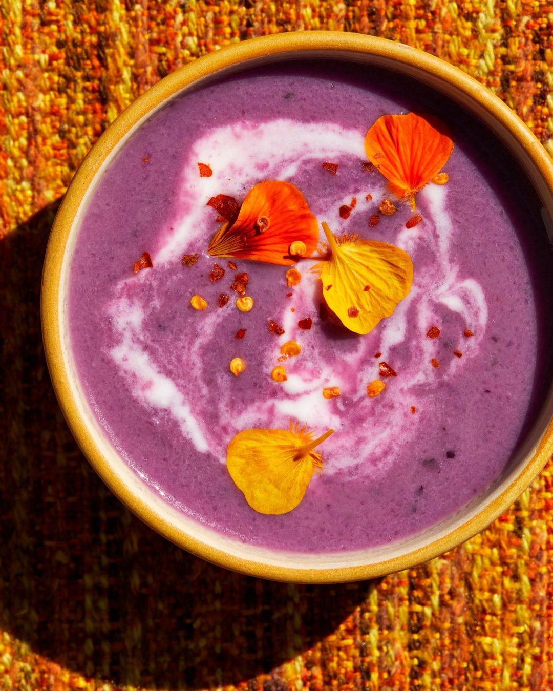 Overhead shot of vibrant purple soup in a tan bowl, garnished with edible orange and yellow flowers and red pepper flakes.