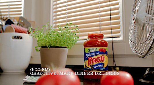 Close-up on a jar of Ragu pasta sauce, potted plant, kitchen utensils in a white holder, and tomatoes in an indoor kitchen setting.